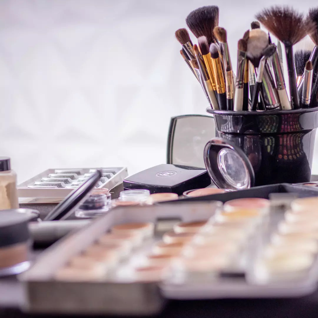 Verschillende make-up producten waaronder een standaard met 15 make up borstels, een oogschaduw pallet, een bronzer en meerdere nagellak potjes. Allemaal geplaatst op een witte tafel. 