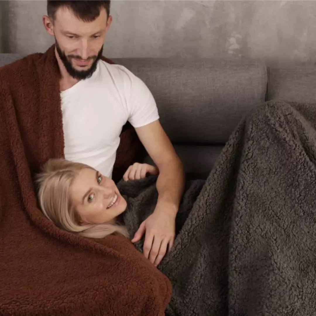 De Love Blanket Liefdesdeken in het grijs en bruin. Een koppel ligt op de bank met onze comfortabele Love Blanket Nederland - zacht, wasbaar en waterdicht.