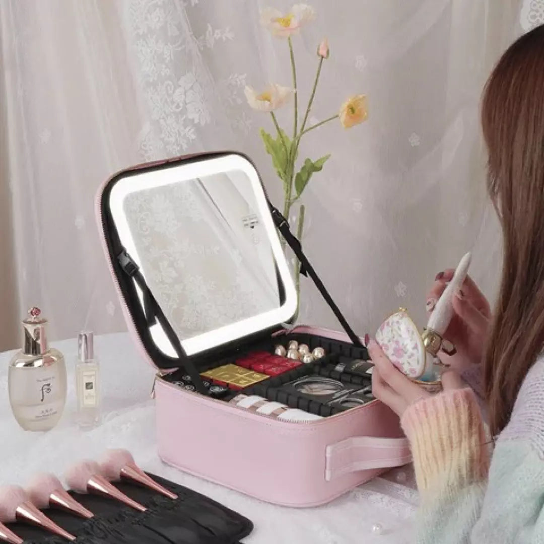 De roze variant van de beste make-up koffer voor thuis en op reis wordt afgebeeld. Een dame zit voor de make up koffer met make up spiegel haar make up te doen. Ze kijkt in de spiegel met verlichting en naast de make up koffer liggen 5 make up borstels.